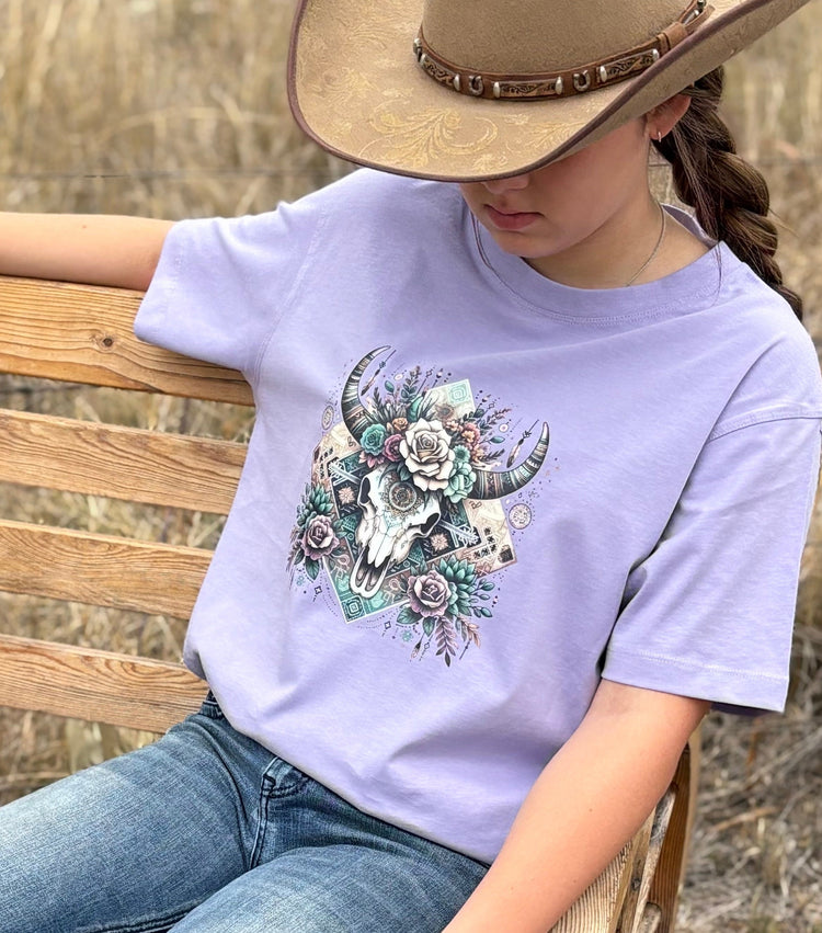 Woman in a lavender Dusty Diamonds bull skull tee and cowboy hat sitting on a timber bench
